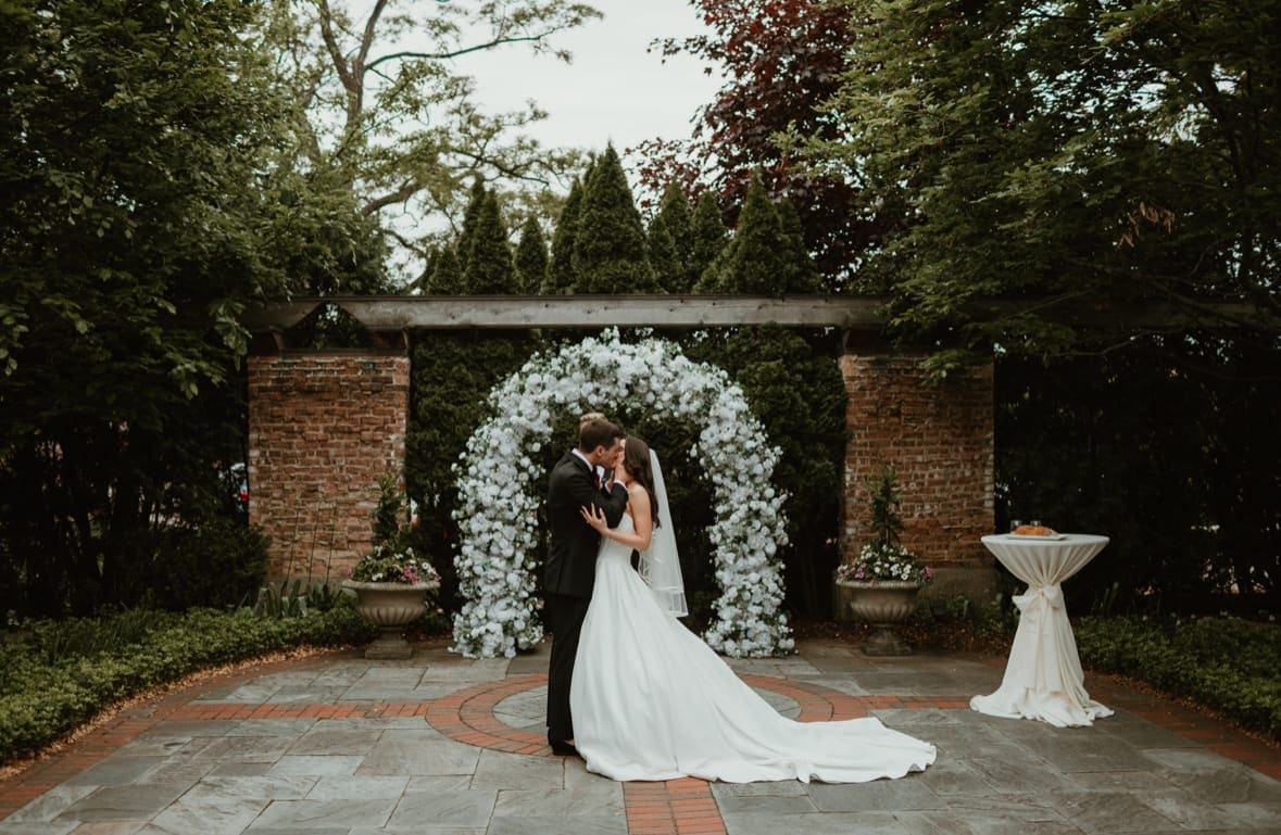 Wedding arch rental Chicago — bride and groom first kiss under white flower arch outdoor garden ceremony